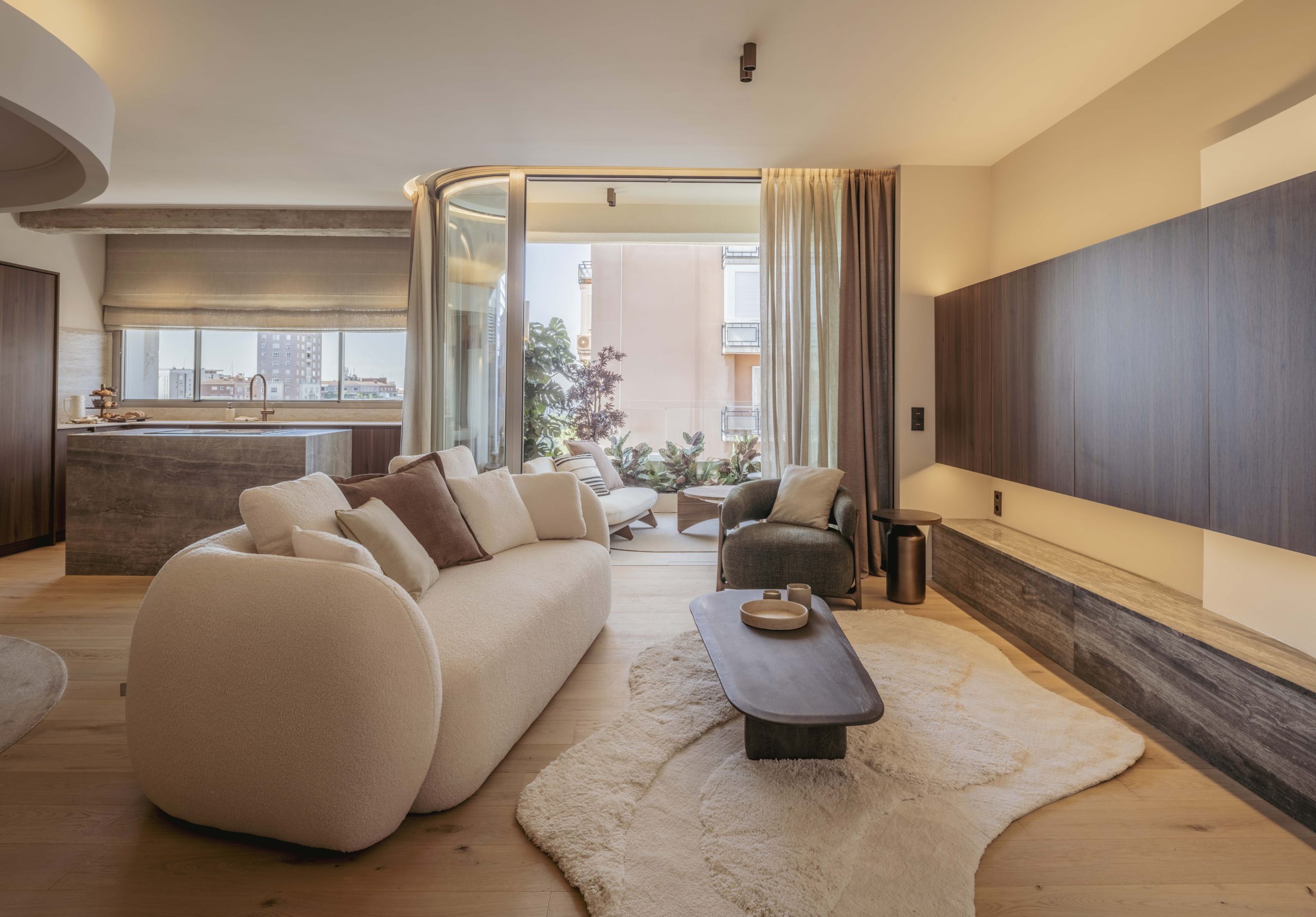 Salón moderno con sofá curvo en tonos claros, cocina abierta y acceso a terraza ajardinada